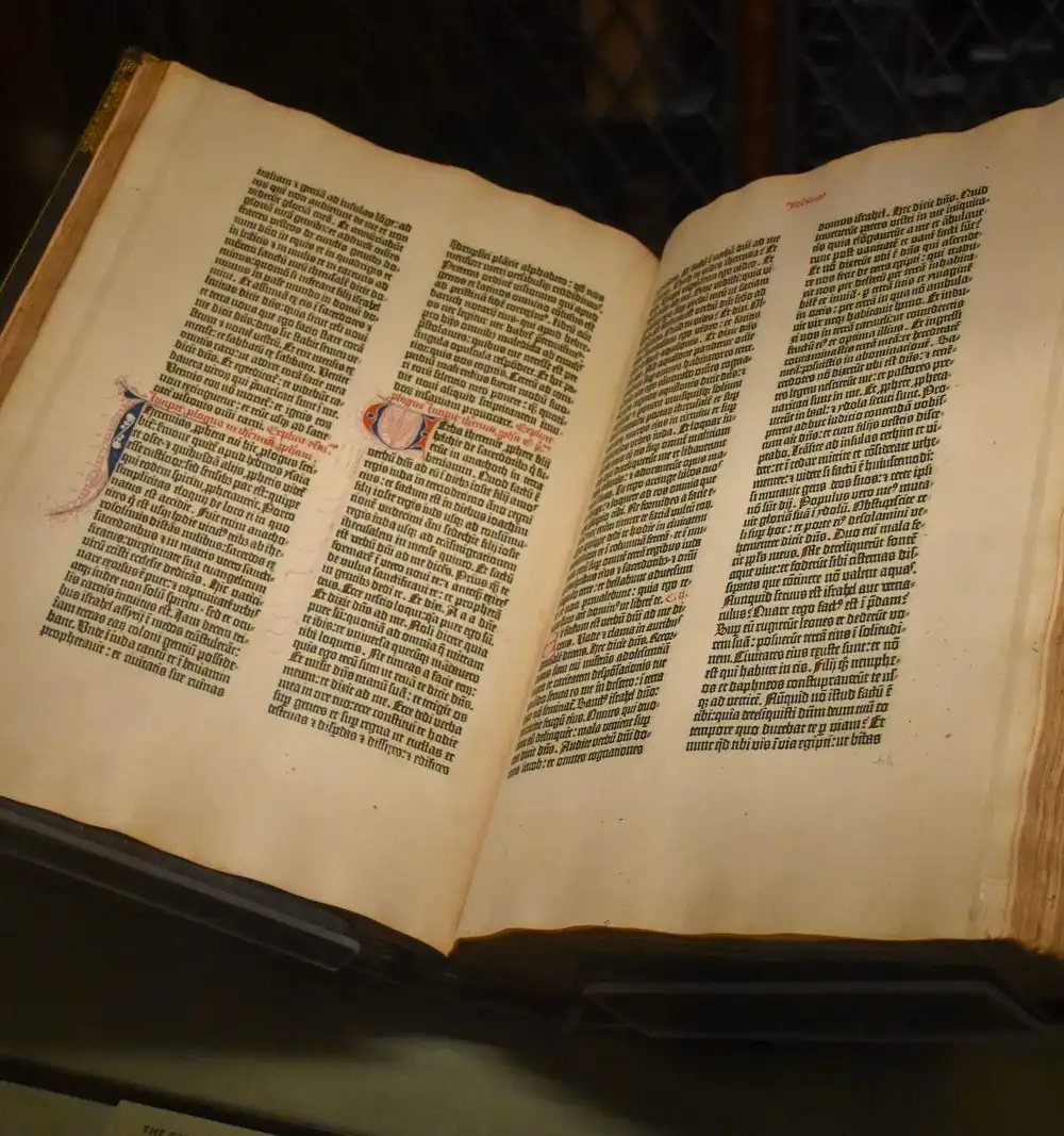 An early printed book lying open in a dark museum case, two dense columns of black text on each page