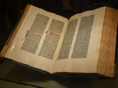 An early printed book lying open in a dark museum case, two dense columns of black text on each page