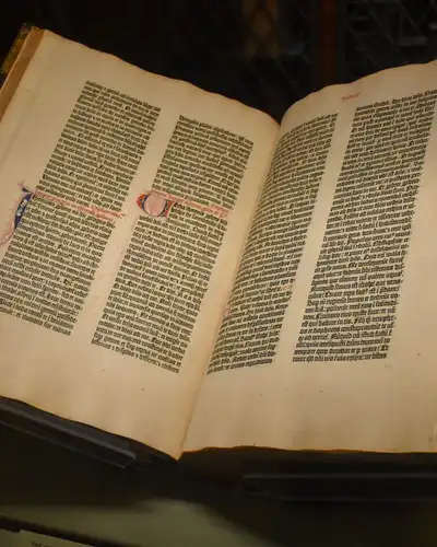 An early printed book lying open in a dark museum case, two dense columns of black text on each page