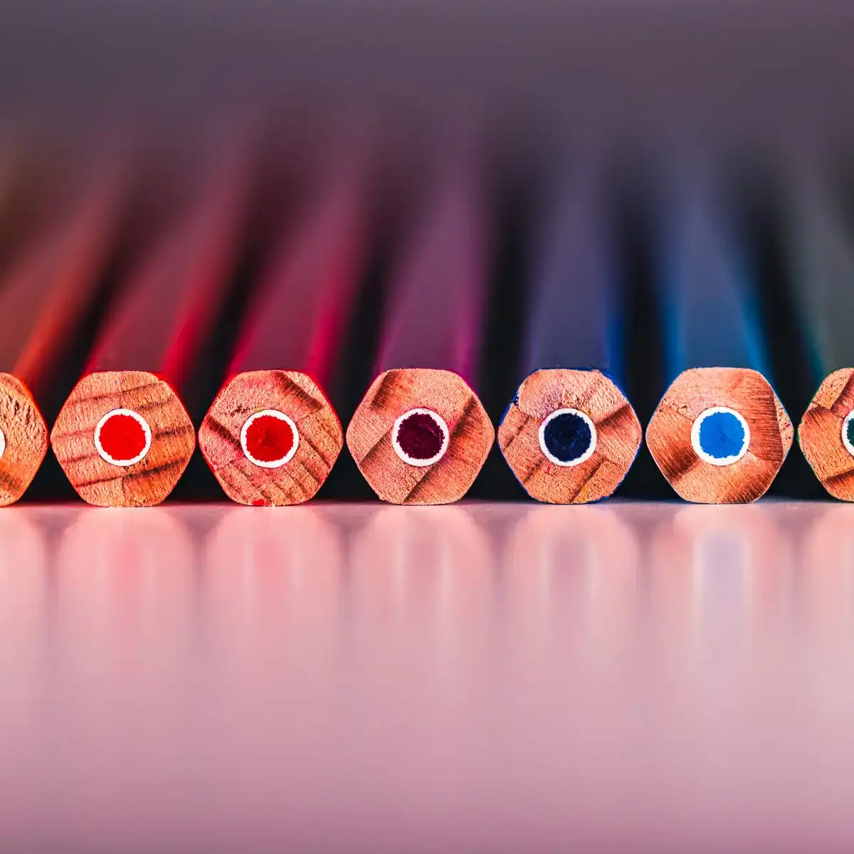 The sharpened ends of nine colored pencils in a row on a pale reflective surface
