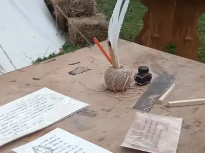 A wooden table outdoors with a quill in an ink pot, a ball of string and handwritten sheets of paper