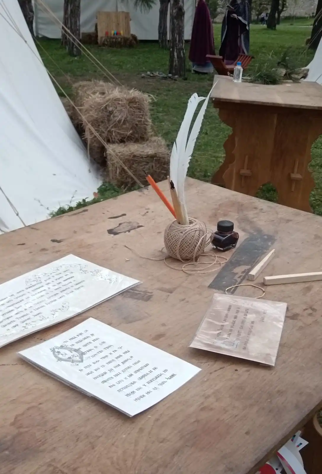 A wooden table outdoors with a quill in an ink pot, a ball of string and handwritten sheets of paper