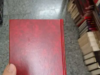 A hand holding a red cloth-bound hardcover book in a bookshop aisle
