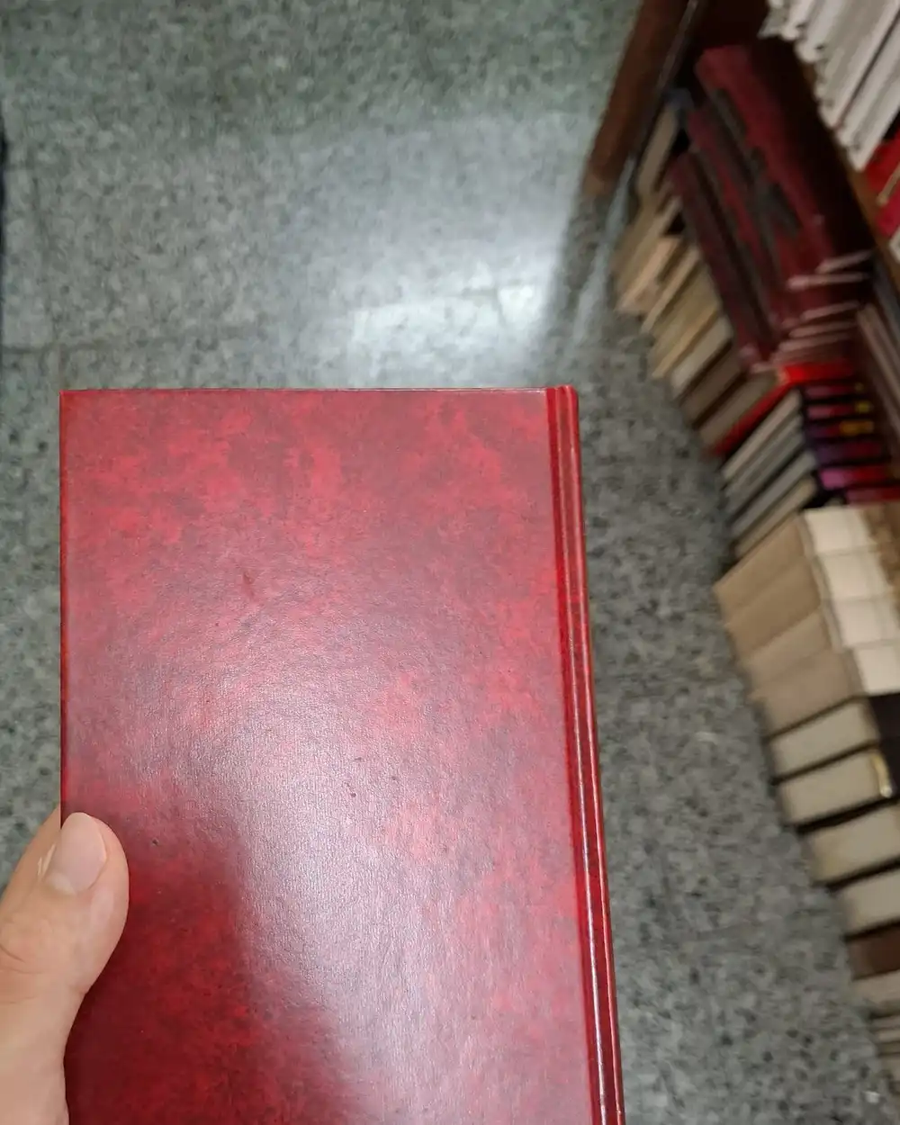 A hand holding a red cloth-bound hardcover book in a bookshop aisle