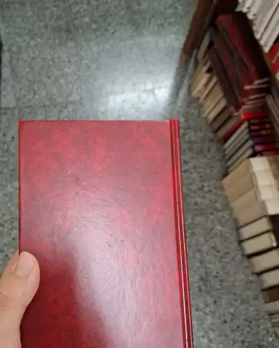 A hand holding a red cloth-bound hardcover book in a bookshop aisle
